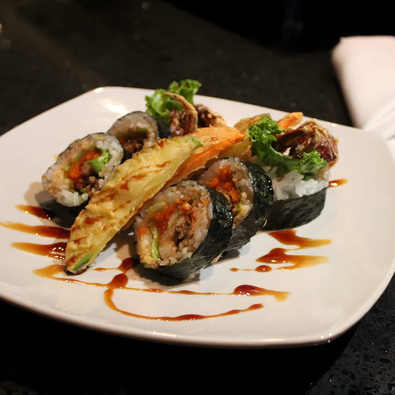 Uni Sushi plate with various sushi rolls, tempura, and soft-shell crab, drizzled with sauce.