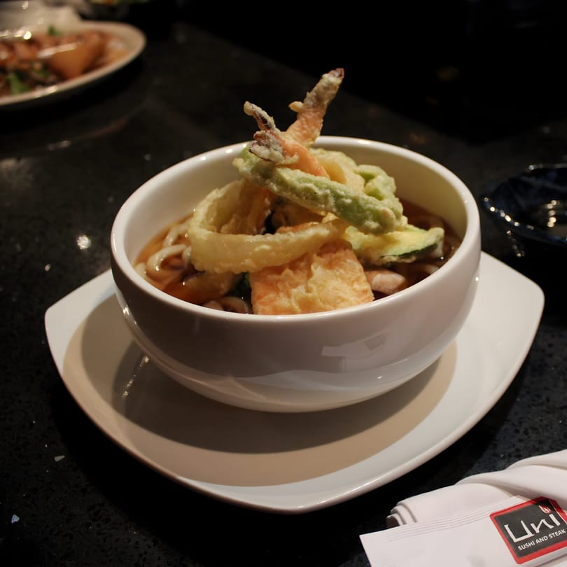A white bowl of Uni Sushi's tempura udon soup with shrimp and vegetable tempura on a dark table.